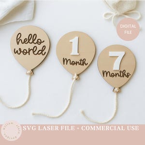 Op de afbeelding: Houten mijlpaalmarkeringen in de vorm van ballonnen. Eén marker zegt "hello world", een andere heeft het nummer "1" en het woord "Month", en de derde heeft het nummer "7" en het woord "Months". Elke ballon heeft een touwtje.