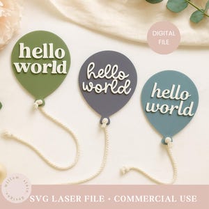Op de afbeelding: Drie ballonvormige decoraties in groen, grijs en blauw, elk met de woorden "hello world" in het wit. Elke ballon heeft een wit touwtje. De afbeelding bevat ook de tekst "DIGITAL FILE" en "SVG LASER FILE + COMMERCIAL USE".