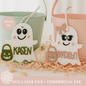 Op de afbeelding: Twee witte spookvormige Halloween-ornamenten met zwarte ogen en glimlachjes, elk met een pompoenvormige traktatiezak. Eén heeft de naam "KASEN" en de andere "MADISON". De ornamenten hangen voor pastelkleurige emmers.