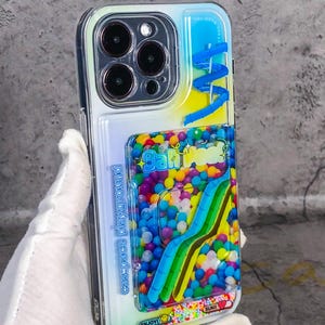 Puede incluir: Una funda transparente para teléfono con un diseño colorido y brillante. La funda tiene un porta tarjetas integrado con un arcoíris de pequeñas bolas en su interior. La funda también tiene el texto "space playground" y "enjoy!" impreso en ella.
