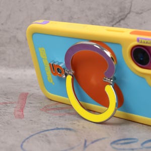 May include: A yellow and blue phone case with a pop-up phone grip. The grip is a yellow and silver hoop attached to a purple and orange design. The phone case has the text "by star" in silver.