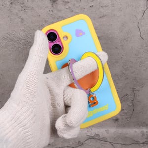 May include: A yellow and blue phone case with a ring holder. The case has a light blue background with a small purple graphic. The ring holder is purple, yellow, and orange with a small charm that says "g" with a gem.