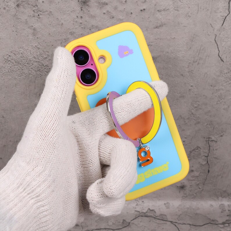 May include: A yellow and blue phone case with a ring holder. The case has a light blue background with a small purple graphic. The ring holder is purple, yellow, and orange with a small charm that says "g" with a gem.