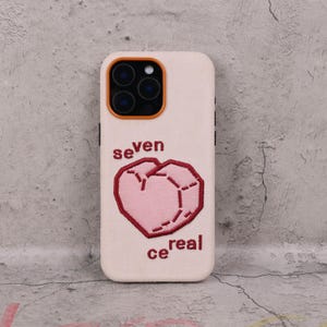 Op de afbeelding: Een lichtgekleurde telefoonhoes met een roze geborduurd hartontwerp en de woorden "seven cereal" in het rood. De hoes heeft een camera-uitsparing met oranje accenten.