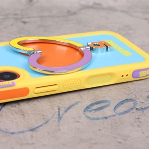 May include: A yellow phone case with a heart-shaped phone grip. The grip is yellow, orange, and purple. The phone case has a light blue back and orange accents. The case has a camera cutout and volume buttons.