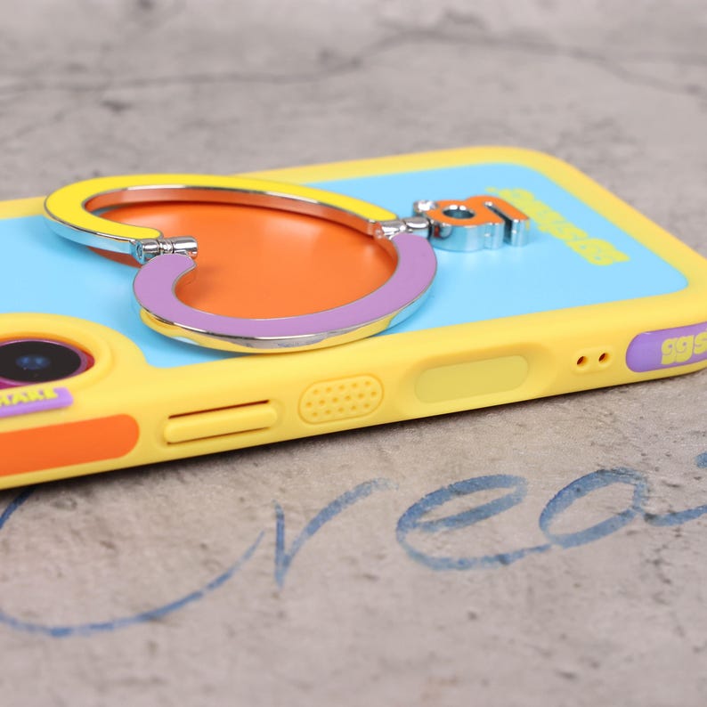 May include: A yellow phone case with a heart-shaped phone grip. The grip is yellow, orange, and purple. The phone case has a light blue back and orange accents. The case has a camera cutout and volume buttons.