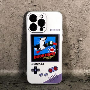 May include: White iPhone case with a Game Boy inspired design featuring the Duck Hunt game. The case has a white background with a gray Game Boy screen showing the Duck Hunt game. The case also has a gray Game Boy button layout with a red and white start button.