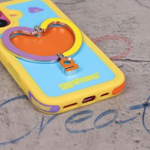 May include: A yellow, blue, and orange phone case with a heart-shaped grip. The case has a blue background with the text "gg share" and a heart-shaped grip with a silver accent. The case is designed to protect the phone.