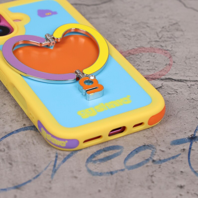May include: A yellow, blue, and orange phone case with a heart-shaped grip. The case has a blue background with the text "gg share" and a heart-shaped grip with a silver accent. The case is designed to protect the phone.