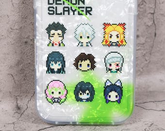 May include: Clear phone case with a pixelated design featuring characters from "Demon Slayer." The case has a pearlescent background with a green and white gradient at the bottom. The text "DEMON SLAYER" is at the top.