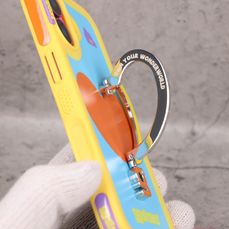 May include: A yellow phone case with a blue and orange design and a silver ring holder. The ring holder is inscribed with the words "Own your Wonder World". The case has a colorful, playful aesthetic.