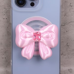 Peut inclure: Un support de téléphone rose clair avec un motif de nœud orné d'une gemme rose rectangulaire au centre. Le nœud a une texture satinée et est fixé à une base circulaire transparente. L'étui du téléphone est bleu clair.