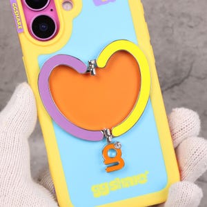 May include: A yellow and blue phone case with a heart-shaped phone grip. The grip is outlined in purple and yellow, with an orange center. The grip has a dangling orange charm with the letter "g" and a small gem. The text "ggshare" is visible.