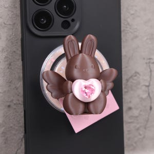 May include: A brown bunny phone grip with wings and a pink heart-shaped gem. The bunny is attached to a black phone case. A pink square is beneath the bunny.