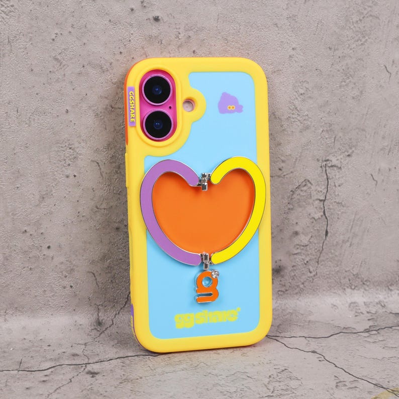 May include: A yellow, blue, and orange phone case with a heart-shaped design. The case has a yellow border, a blue background, and an orange heart in the center. The case has the text "GGSHARE" and a small purple and yellow heart charm.