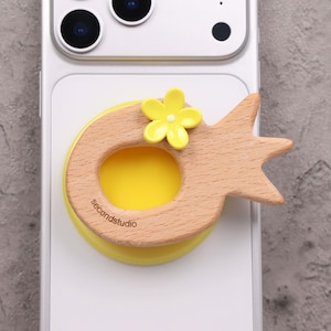 May include: A wooden phone grip with a yellow flower and a yellow base is attached to a white phone. The grip has the text "secondstudio" on it. The grip is shaped like a stylized bird.
