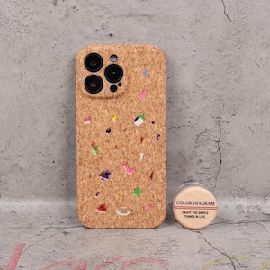 Puede incluir: Una funda de teléfono hecha de corcho con motas de colores. La funda tiene un recorte para la cámara y un pequeño recipiente redondo con el texto "COLOR DIAGRAM" y "ENJOY THE SIMPLE THINGS IN LIFE."