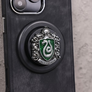 May include: A black phone case with a silver and green Slytherin crest phone grip. The crest features a snake on a green background with the word "Slytherin" below. The phone case has a textured appearance.
