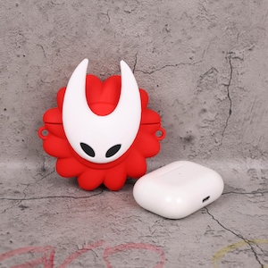 May include: A red and white silicone case for wireless earbuds, featuring a stylized character design. The case is shaped like a flower with a white face and horns. A white wireless earbud case is next to it.