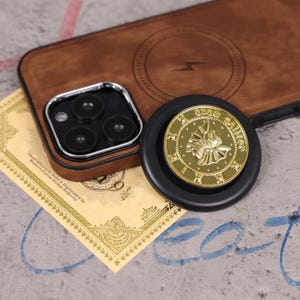 Puede incluir: Una funda de teléfono de cuero marrón con un protector de lente de cámara plateado y una moneda de oro sobre una base negra. La moneda tiene un diseño en relieve y texto. Un documento amarillo es parcialmente visible.
