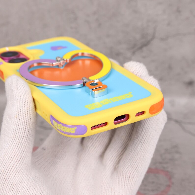 May include: Yellow, blue, and orange phone case with a heart-shaped ring holder. The case has the text "Be Shower" and "Wishare". The ring holder is silver, purple, and yellow. The phone case is held by a person wearing a white glove.