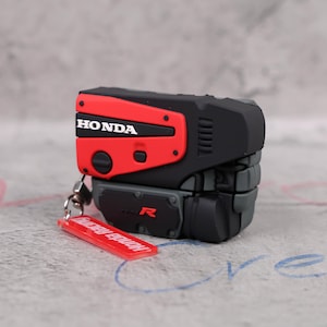 May include: A black and red Honda engine-shaped keyring. The keyring features the word "HONDA" in white lettering on a red panel. A small, red rectangular tag with "Honda Racing" is attached.