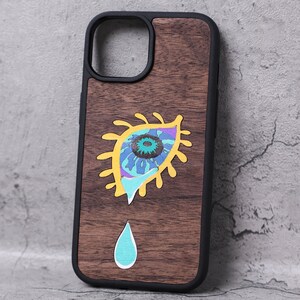 以下が含まれることがあります： 黒い縁取りの木製スマホケース。黄色、青、紫のアクセントと、ターコイズの涙が特徴的なカラフルな目のデザイン。スマートフォン向けに設計されています。