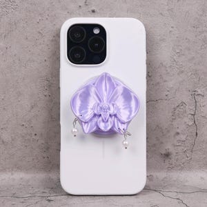 Puede incluir: Funda de teléfono blanca con un agarre para teléfono en forma de orquídea lavanda. La orquídea tiene un acabado satinado y está adornada con colgantes de perlas de imitación. El teléfono tiene una cámara de triple lente.