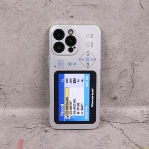 以下が含まれることがあります： カメラのようなデザインのシルバーの携帯電話ケース。複数のレンズ、ボタン、カメラ設定を表示する画面があります。画面には、ホワイトバランス、感度、アスペクト比、画像サイズ、品質のオプションが表示されます。「Vavenue」のブランド名が表示されています。