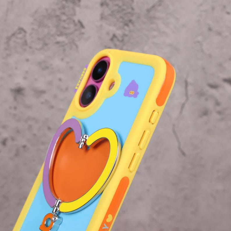 May include: A colorful phone case with a heart-shaped grip. The case features a yellow border, a light blue center, and an orange heart with purple and yellow accents. A small charm hangs from the heart.