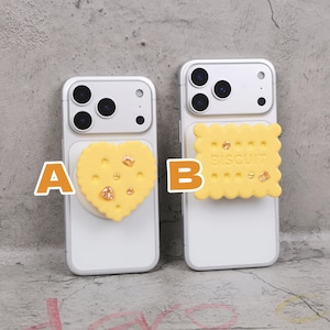 May include: Two white cell phone cases with decorative yellow biscuit-shaped attachments. One is heart-shaped and the other is a square biscuit with the word "BISCUIT" printed on it. Both have small, sparkling embellishments.
