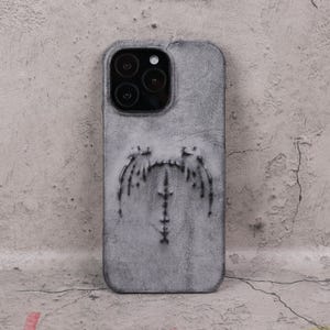 Może przedstawiać: Szare skórzane etui na telefon z teksturowanym wykończeniem. Etui ma stylizowany wzór na plecach, z centralną pionową linią i kształtami przypominającymi skrzydła. Obiektywy aparatu są widoczne u góry.