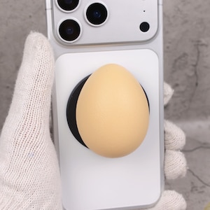 May include: A light beige egg-shaped phone grip attached to a white phone case. The grip is mounted on a black circular base. The phone case is attached to a silver phone.