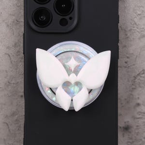 May include: White butterfly-shaped phone grip attached to a black phone case. The grip has a pearlescent background with a four-pointed star design. The butterfly has a heart-shaped center.