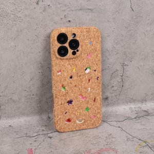 May include: A light brown phone case made of cork material. The case has a speckled design with colorful flecks of pink, yellow, blue, and green. The case has a camera cutout with three lenses.