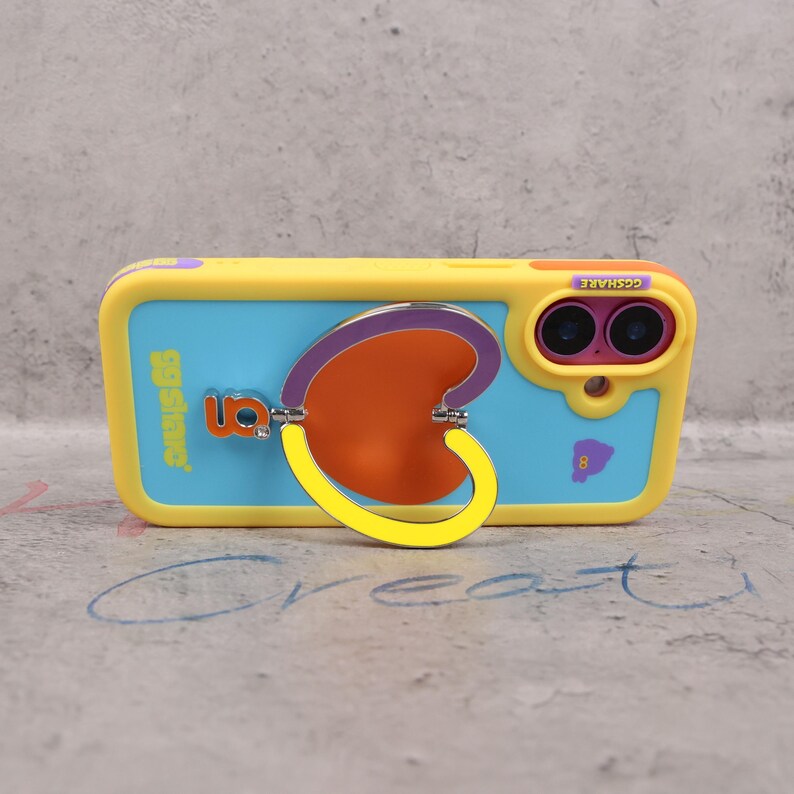 May include: A yellow and blue phone case with a heart-shaped ring holder. The case has the text "ggshare" and "8" printed on it. The ring holder is orange, purple, and yellow.