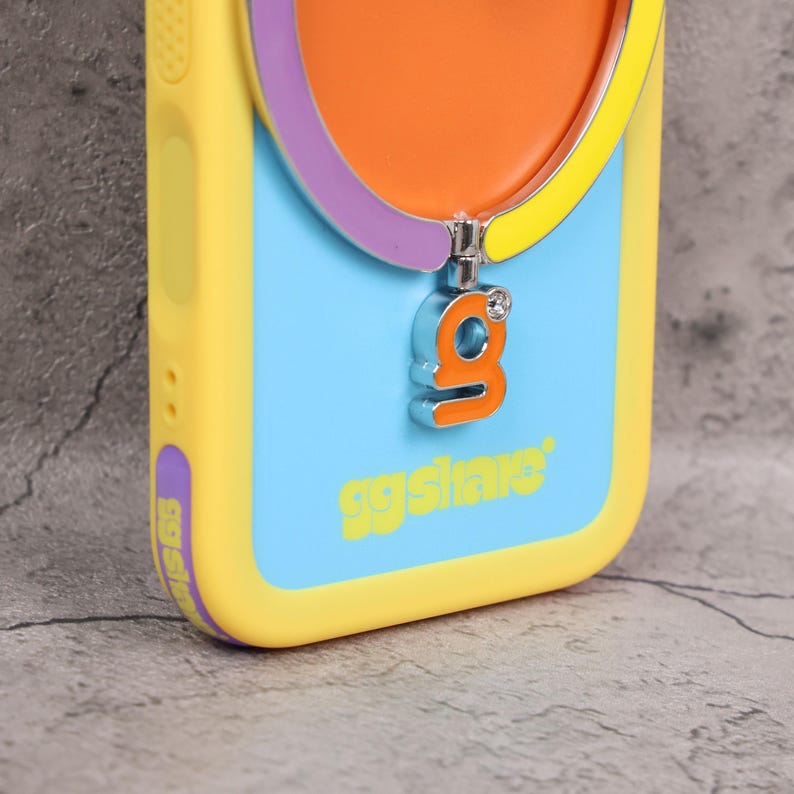 May include: A yellow, blue, and orange phone case with a silver charm. The charm is in the shape of the letter "g" with a small gem. The case has the text "ggslive" in yellow.