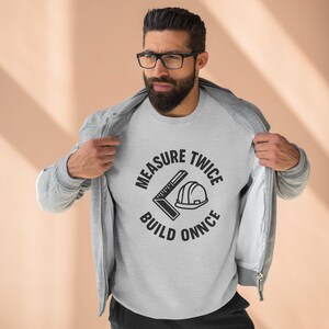 May include: A light gray sweatshirt with the text "MEASURE TWICE BUILD ONNCE" in a circular design. The design includes a square ruler and a hard hat. The person is wearing a gray zip-up hoodie.