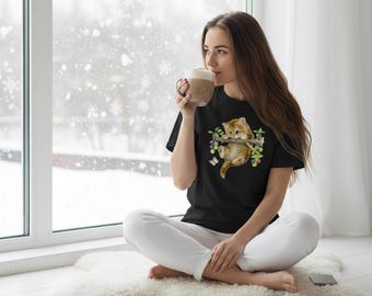 Puede incluir: Camiseta negra con una ilustración de acuarela de dos gatitos naranjas en una rama con bayas y mariposas. Los gatitos son el punto focal del diseño. La persona está sentada en una alfombra blanca cerca de una ventana.