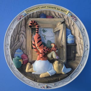 Könnte beinhalten: Eine dekorative runde Wandplatte mit einer Szene mit Winnie Puuh und Tigger. Tigger, mit orangefarbenen und schwarzen Streifen, hüpft auf Puuh. Die Platte trägt den Text "Bouncing's what Tiggers do best".
