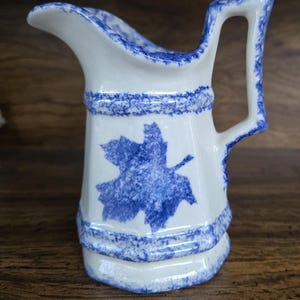 May include: A white ceramic creamer with a blue and white maple leaf design. The creamer has a handle and a spout.