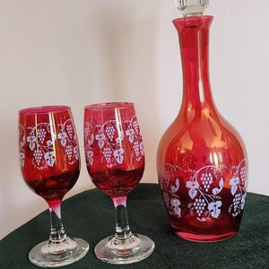 May include: A set of ruby red glassware, including a decanter with a clear stopper and two wine glasses. Each piece features a white grape and leaf pattern. The items are displayed on a dark green surface.