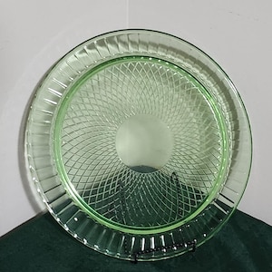 May include: A vintage green glass plate with a textured, crisscross pattern in the center and a ribbed border. The plate is round and sits on a black metal stand, showcasing its design. This decorative plate is a classic piece.