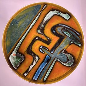 May include: A round decorative plate with an abstract design. The plate features a central orange area with black, blue, and green shapes. A green and blue section forms a border. The plate has a white rim.