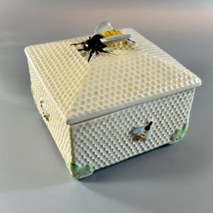 May include: A white ceramic box with a honeycomb pattern and a bee on the lid. The box has a bee on each side and green leaves on the bottom.