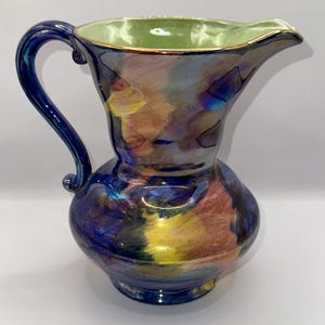 Puede incluir: Una jarra decorativa de cerámica con base bulbosa y asa curva. La jarra presenta un esmalte iridiscente vibrante con tonos de azul, amarillo y rosa. El interior es verde claro con un borde dorado. La jarra mide aproximadamente 25 cm de altura.
