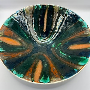 Puede incluir: Un cuenco decorativo de cerámica con borde plateado. El cuenco presenta un patrón arremolinado de colores verde oscuro, naranja y marrón. El centro del cuenco es azul oscuro, y el diseño general es abstracto y artístico.