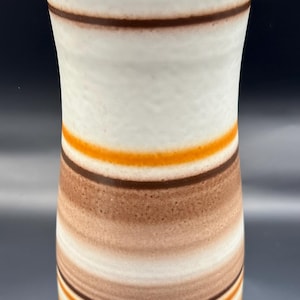 Könnte beinhalten: Eine weiße Keramikvase mit horizontalen Bändern in Orange, Braun und Beige. Die Vase hat eine leicht ausgestellte Form und eine glatte, glänzende Oberfläche. Das Design ist im Retro-Stil.