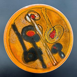 Puede incluir: Un plato redondo de cerámica naranja con una superficie texturizada. El plato presenta un diseño abstracto negro, rojo y amarillo. El diseño incluye formas que se asemejan a instrumentos musicales. El plato tiene un borde blanco.