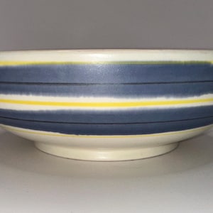 May include: A ceramic bowl with a cream base and a wide, rounded shape. The bowl has horizontal stripes in navy blue and yellow, creating a decorative pattern. The bowl is suitable for serving food.